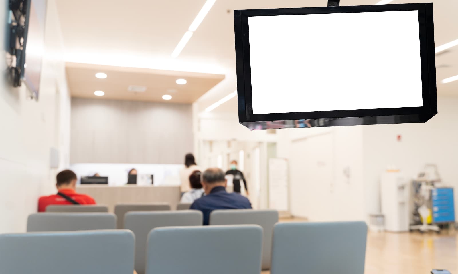 Waiting room with seated people and a blank digital display screen overhead.
