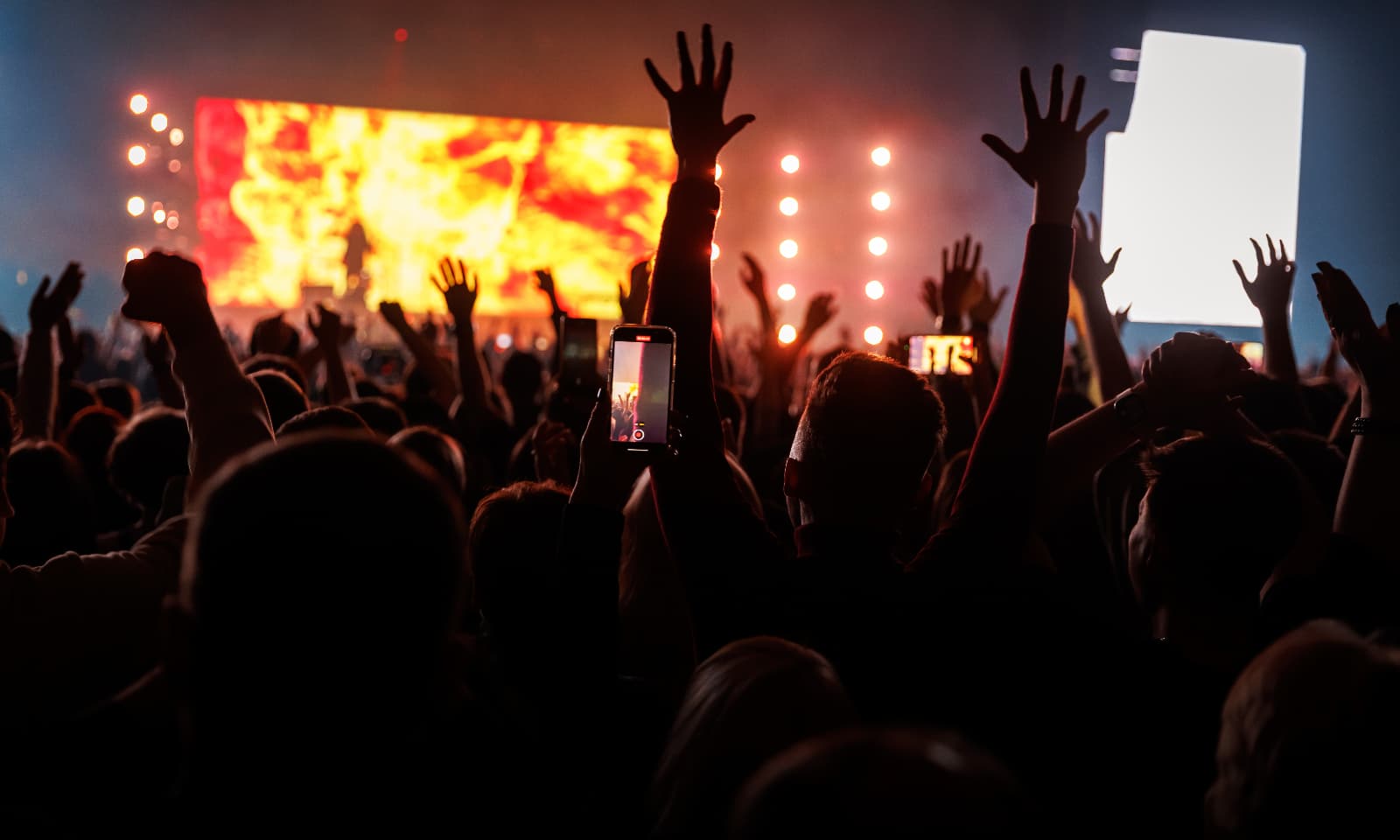 Crowd at a live music concert with raised hands, bright stage lighting and large LED screens in the background