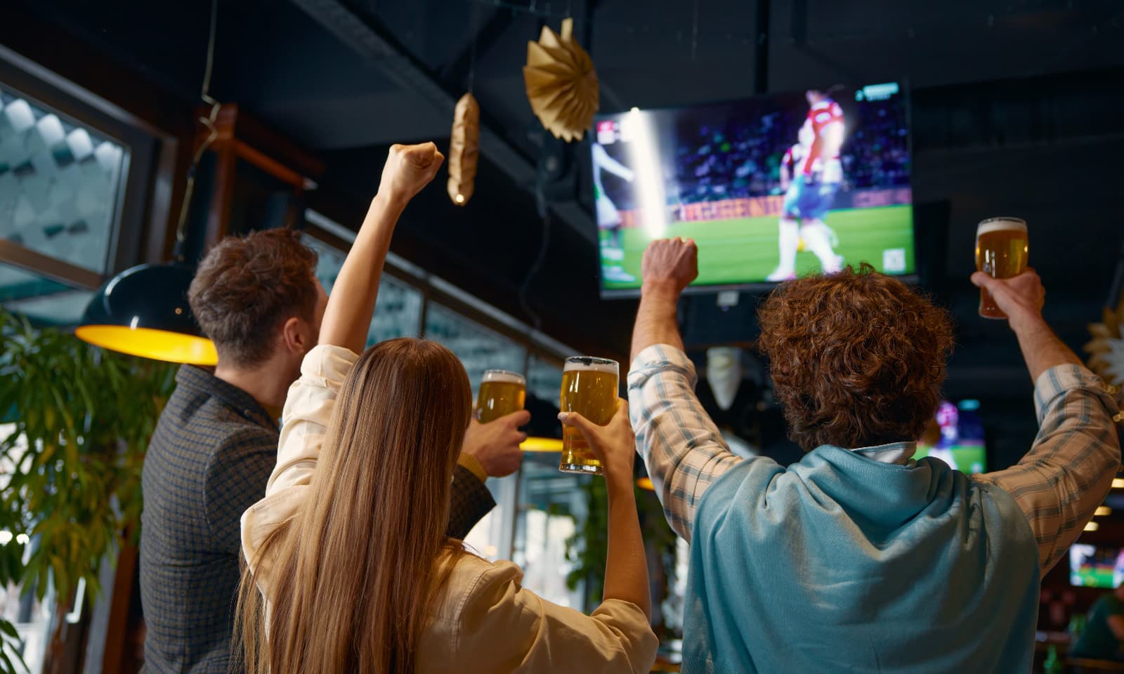 Group of friends cheering and holding drinks while watching a football match on a TV screen in a pub